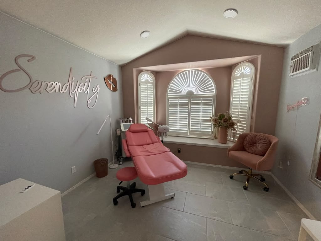 The full treatment room at Serendipity in El Paso — the Serendipity script on the wall, a rose treatment chair, an arched shuttered window with flowers, and a velvet chair.